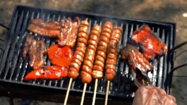 El Barbekü Izgara Et Için Mutfak Tongs kullanma - Rack Focus. Izgara Et ve Sebze. Izgara Hot Dog Sosis, Pastırma ve Kırmızı Biber Bir Barbekü Üzerinde Kömürler Over - Yavaş Hareket.