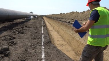 İnşaat Sahasında Petrokimyasal Gaz Boru Hattı Mühendisi Kontrol. Mühendis Kırmızı Hardhat giyen ve Pano ya yazma. Borular siper yanında destekleyici kum torbaları üzerine serilir ve birlikte kaynaklanır.