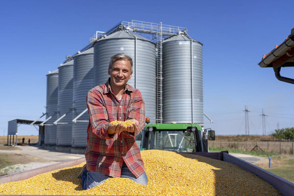 Smiling Farmer Sitting in Tractor Trailer Full of Corn Seeds in  front of Farm Grain Bins
