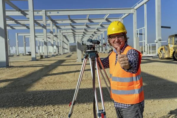 Glimlachende bouwvakker met Tacheometer die duim opgeeft op een bouwplaats — Stockfoto