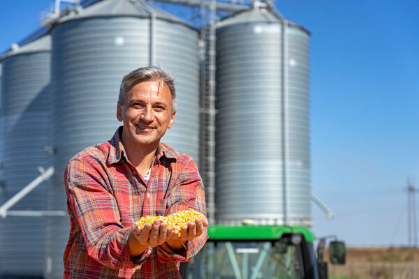 Smiling Farmer Showing Freshly Harvested Corn Grains Against Grain Silo. Farmer's Hands Holding Harvested Grain Corn. Farmer with corn kernels in his hands sitting in trailer full of corn seeds.