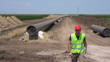 İnşaat Ustası Pipeline İnşaat Alanında Panoya Yazıyor. Kırmızı Şapkalı ve Sarı Yelekli Bir İşçinin Portresi. Mühendis Petrol veya Gaz Boru Hattı 'nın yanında duruyor.