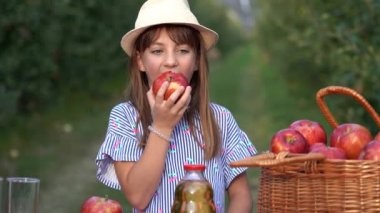 Güneşli Meyve Bahçesinde Kırmızı Elma Yiyen Şirin Mavi Gözlü Kız. Güzel Gözlü Gülümseyen Küçük Kız Kameraya Bakıyor. Sağlıklı gıda konsepti. Şapkalı ve Taze Elmalı Şirin Kız Fotoğrafı.