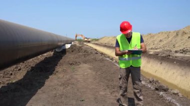 İnşaat Ustası Gaz Boru Hattı İnşaat Alanında Radyo İletişim Cihazı ile konuşuyor. Kırmızı Şapkalı ve Sarı Yelekli Bir İşçinin Portresi. Mühendis Petrol veya Gaz Boru Hattı 'nın yanında duruyor.