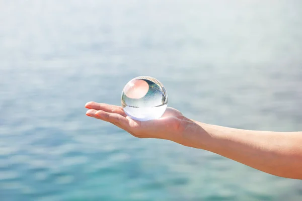 A female hand holds a glass ball with the reflection of the sea and sky. Concept of summer holidays and vacation trips. Can illustrate the opening of the beach season.