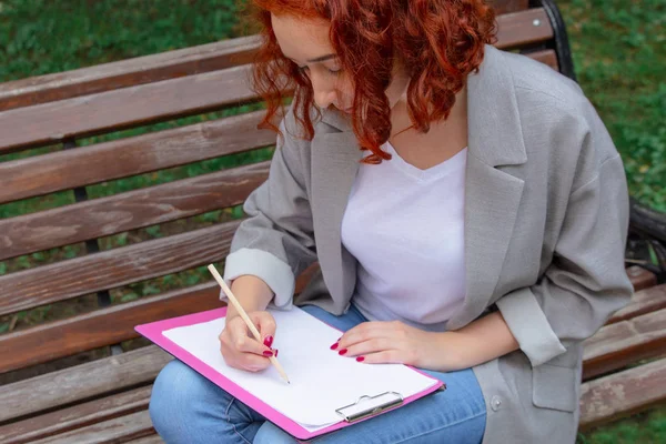 ベンチに市内の公園に赤い髪の少女は 紙のためのタブレットの鉛筆を描画します 女の子は赤髪 — ストック写真