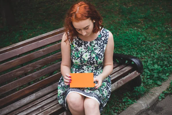 赤い髪の少女が公園のベンチにはがきを読み取ります — ストック写真