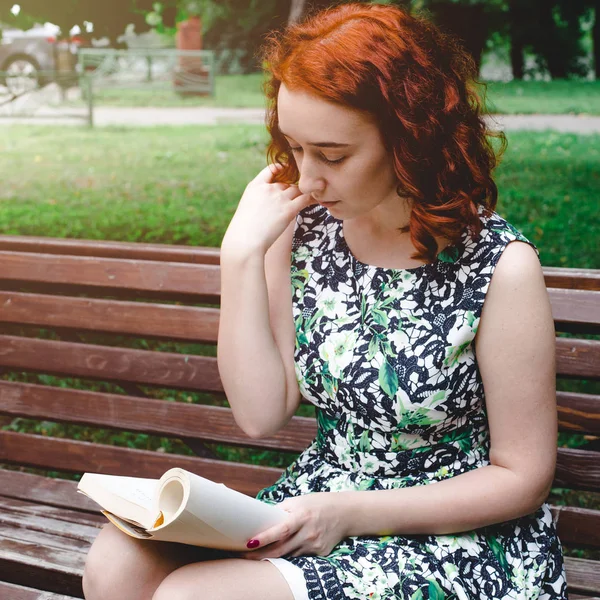 一个红头发的女孩正在公园长椅上看书 — 图库照片