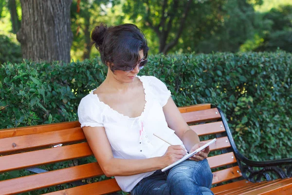 一个女孩在公园长椅上的笔记本上写字 身穿蓝色牛仔裤和白色上衣的中年黑发妇女 — 图库照片