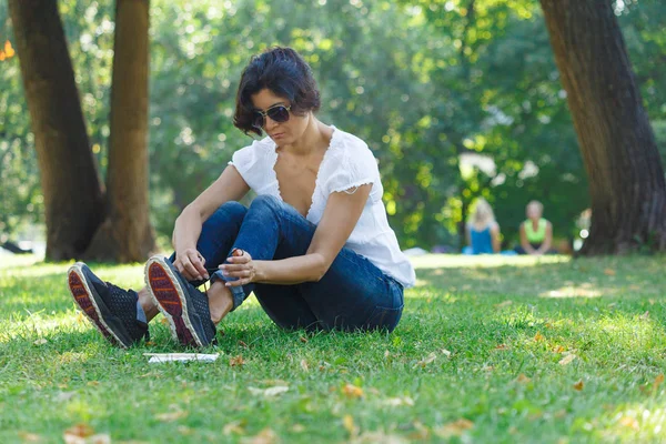 The brunette girl in the park ties up her shoelaces on sneakers. A concept of calm mood and relaxation in a park on the green grass. A middle-aged woman in blue jeans.