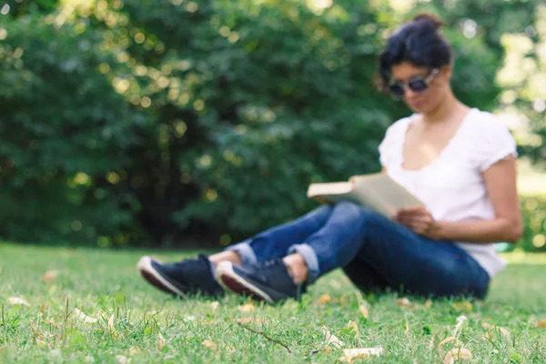 在公园里放松 在草地背景下 一个女孩读了一本书 一个穿着蓝色牛仔裤的黑发女人在柔软的焦点 — 图库照片