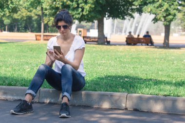 Kaldırıma park bir cep telefonu ile esmer bir kadın oturuyor. Şehir Park rekreasyon kavramı.