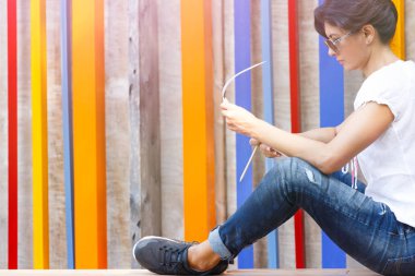 Kot pantolon ve güneş gözlüğü bir esmer bir ahşap sahne alanı'nda oturur ve notları bir defter okur.