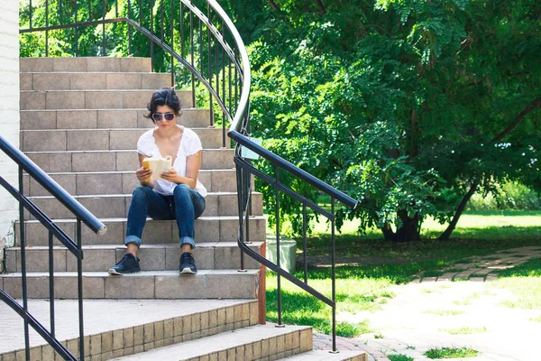 一个女孩坐在台阶上 在公园里看书 那个女人戴着太阳镜 — 图库照片