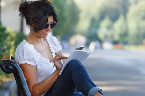 Ein Mädchen Mit Sonnenbrille Zeichnet Einem Notizbuch Auf Einer Parkbank — Stockfoto
