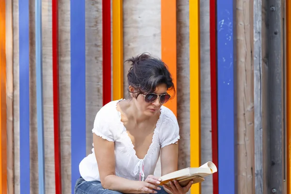 一个戴墨镜的女人坐在木制的舞台上看书 — 图库照片
