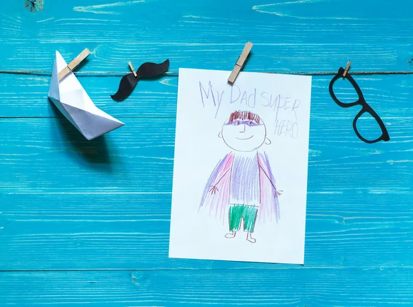 Children\'s drawing with the inscription my dad is a super hero secured with a clothespin with a paper boat and a paper mustache, on a blue background
