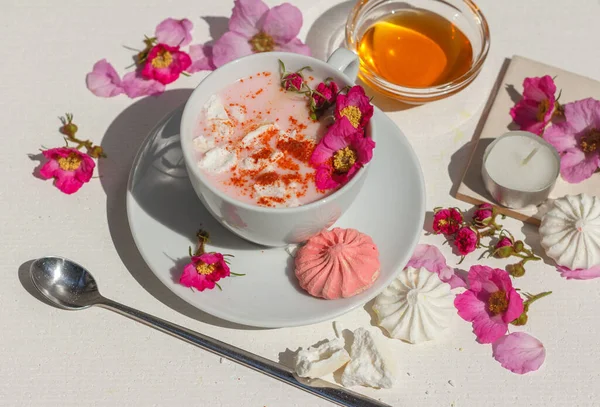 Cup with pink moon milk, with the addition of rosehip flowers, cinnamon, turmeric and other bright spices. On a white table with colored meringues, flowers and honey. Close-up