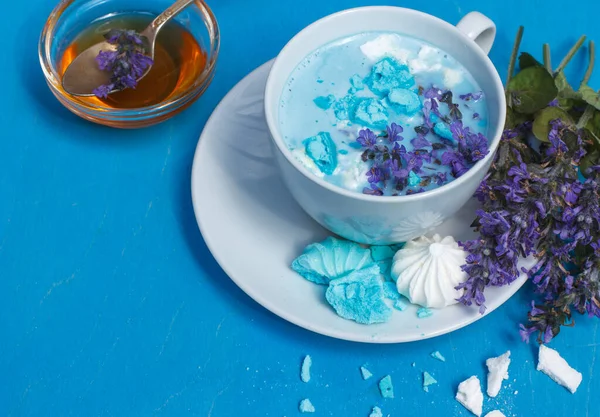 Blue moon milk in a white Cup with honey and blue flowers, decorated with blue and white meringue. On a blue background, close-up