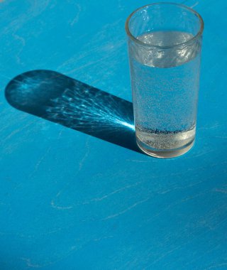 Bir bardak su, güneşin parlak ışıklarıyla aydınlanan, mavi arka planda güzel bir gölgesi olan.