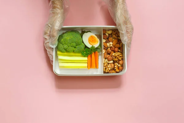 Hands in protective gloves with a healthy lunch in a box. Horizontal top view on a pink background