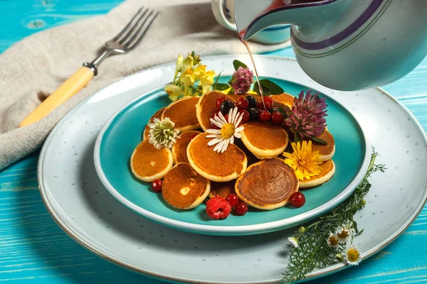 Eine Portion Mini-Pfannkuchen auf einem blauen Teller, dekoriert mit frischen Blumen und einer Kanne mit Gießsirup. Nahaufnahme — Stockfoto