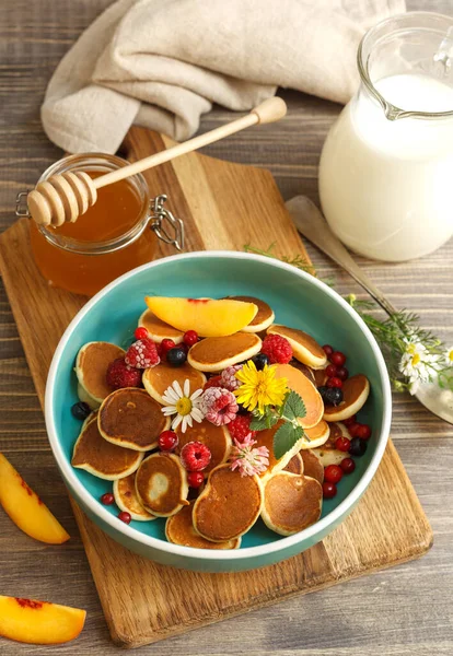 Mini-Pfannkuchen mit frischen Blumen und gefrorenen Beeren in einer blauen Schüssel verziert. Auf einem Holztisch mit einem Krug Milch und Honig im Glas — Stockfoto