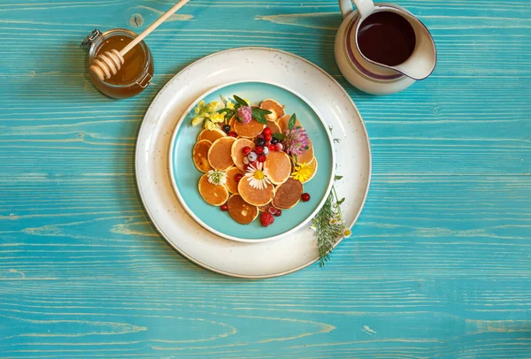 Ein Teller mit leckeren Mini-Pfannkuchen mit gefrorenen Beeren und Blumen. Auf blauem Holzgrund. In der Mitte, mit dem Raum — Stockfoto