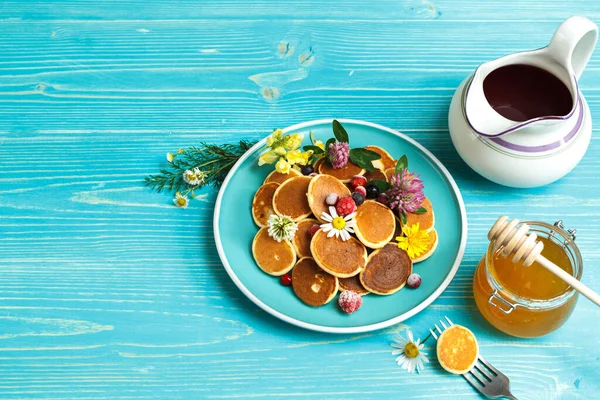 Kleine Pfannkuchen mit Beeren auf einem blauen Teller. Mit einem Krug Sirup und einem Glas Honig. Auf blauem Holzgrund, mit Platz — Stockfoto