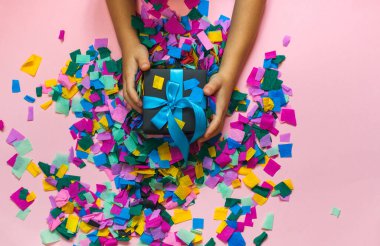 Çocuk mavi kurdeleli bir hediye kutusu tutuyor. Renkli konfetiyle pembe bir yüzeyde. Üst manzara.