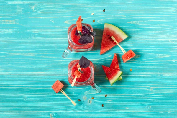 Kırmızı fesleğen yapraklarıyla süslenmiş karpuz smoothie ile iki cam bardak. Mavi ahşap arka planda karpuz dilimleri. Üst