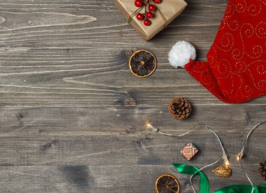 Kırmızı Noel Baba Şapkası, parıldayan çelenk, hediye kutusu, bayram dekorasyonu ve koyu ahşap arka planda kurdeleler. Üst görünüm