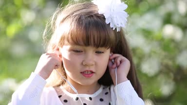 Beyaz elbiseli bir kız küçük ses kulaklık aracılığıyla müzik dinler. Güzel bir kız bir yaz bahçesinde kendini telefondan bir ses kulaklık kulağına koyar.