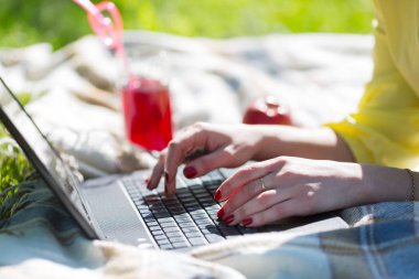 Eller bir piknik dizüstü klavye üzerinde çalışmak. Bir serbest meslek sahibi kız ile bir dizüstü bilgisayar parkta çim üzerinde çalışıyor. Yakınlarda bir bardak meyve suyu ve bir tabak bisküvi ile bulunmaktadır.