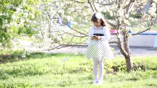 穿着白色连衣裙的女孩通过小音频耳机听音乐 夏日花园里一个美丽的小女孩把自己从电话里的音响耳机里放进耳朵里 — 图库视频影像