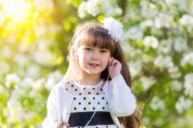 Beyaz elbiseli bir kız küçük ses kulaklık aracılığıyla müzik dinler. Güzel bir kız bir yaz bahçesinde kendini telefondan bir ses kulaklık kulağına koyar.