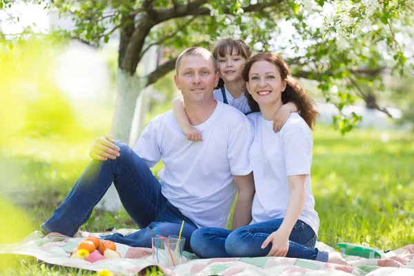 Happy family with a white dog in a summer park. Parents and a 5 year old daughter are sitting in the garden under a blossoming apple tree.