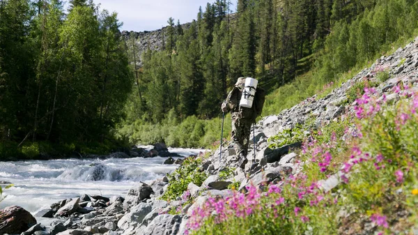 A man with a backpack walks along a mountain river. A tourist with a large bag on his back walks along the rocks near a noisy fast river.