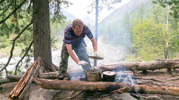 The man is cooking food at the stake in the forest. The tourist interferes with a soup sauce in a saucepan on a ksotra in a hike.