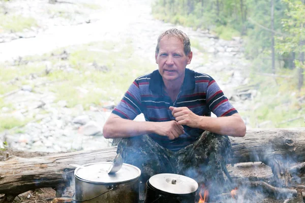 The tourist cooks food at the stake, sits on a fallen tree.