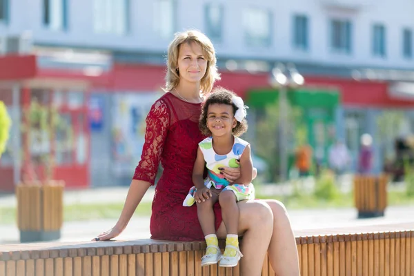 Kaukasische Frau mit schwarzem Mädchen sitzt auf einer Bank. — Stockfoto