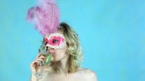一个在嘉年华面具里喝杯酒的女孩 一个戴着羽毛的漂亮面具的金发女郎在蓝色的背景下喝香槟 — 图库视频影像