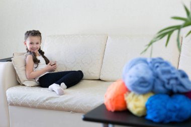 Masanın üzerine konu farklı renklerde örme için vardır. Bir kız kanepede bir cep telefonu ile.