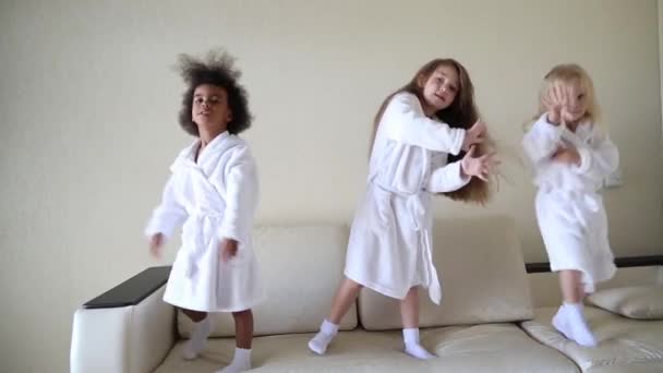 Три Дівчинки Стрибають Дивані Дівчата Різних Національностей Білих Халатах Танцюють — стокове відео