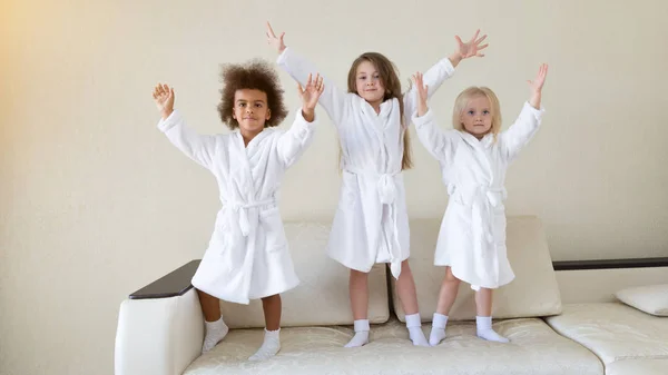 Three little girls dancing on the couch. Girls of different nationalities in white coats dance and laugh after a bath.