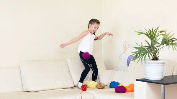 A girl stands on the couch and throws balls of wool yarn up. The child plays with the balls of spools of thread.