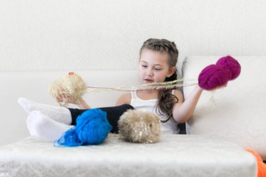 Bir kız ile renkli iplik kanepede oturuyor. Güzel küçük kız bacaklarını masanın üstüne koy ve iplik bobinler.