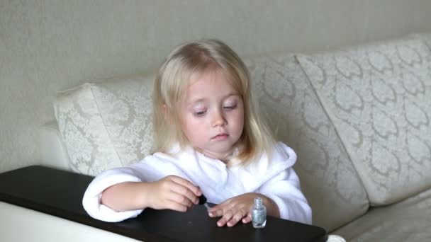 Petite fille caucasienne peint des ongles sur les mains.La fille de 3-4 ans en robe de chambre blanche s'assoit sur le canapé et peint ses ongles avec du vernis .