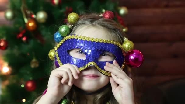 一个戴着狂欢节面具戴着圣诞花环的女孩 一个拿着圣诞球的小女孩在她的头发上玩狂欢节面具 — 图库视频影像