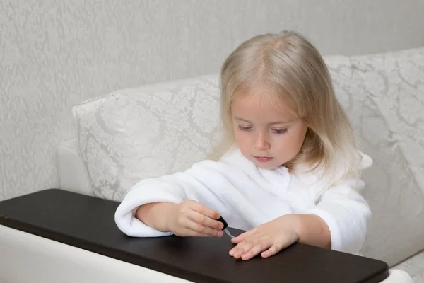 Little Caucasian girl paints nails on hands. The girl of 3-4 years in a white dressing gown sits on the sofa and paints her nails with varnish.